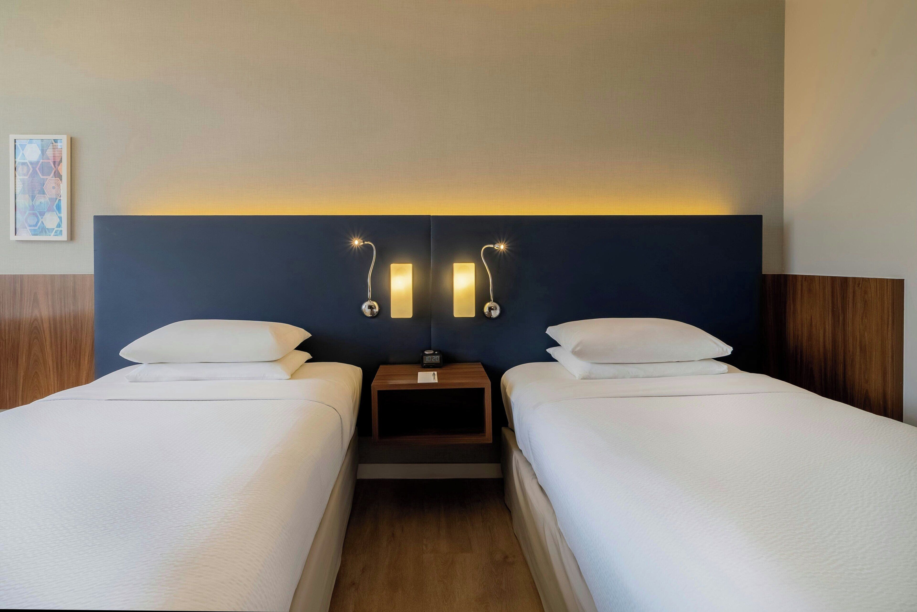 Ein Hotelzimmer mit zwei Einzelbetten, einem Nachttisch mit Uhr und Wandleseleuchten.