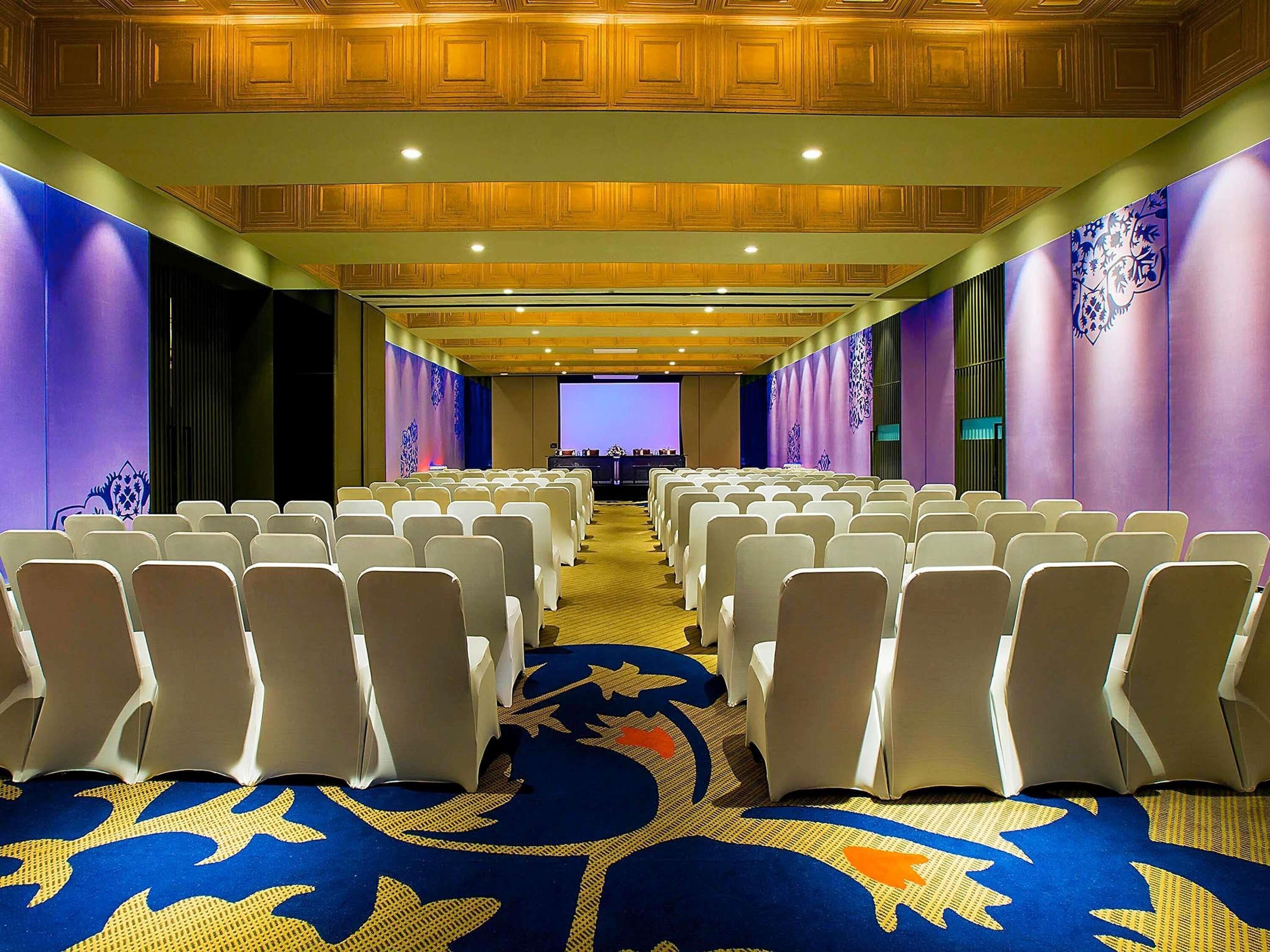A conference room with rows of white chairs, a carpet with a blue and gold pattern, and wooden paneling on the ceiling.