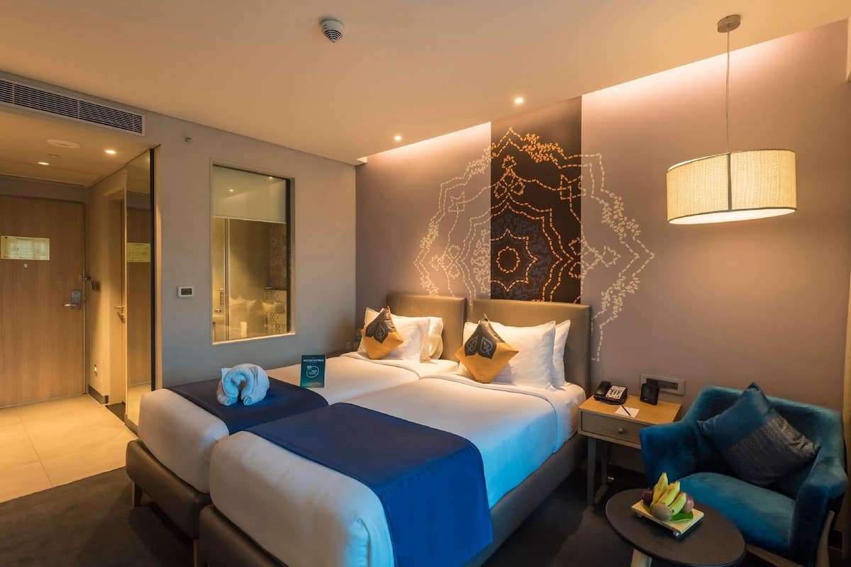 A hotel room with a large bed, a desk with a lamp, a chair, and a small table with fruit.