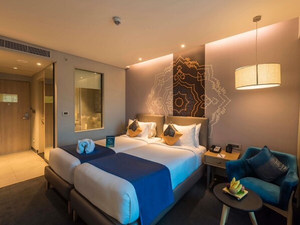 A hotel room with a large bed, a desk with a lamp, a chair, and a small table with fruit. - Grand Mercure Mysore (Mysore)