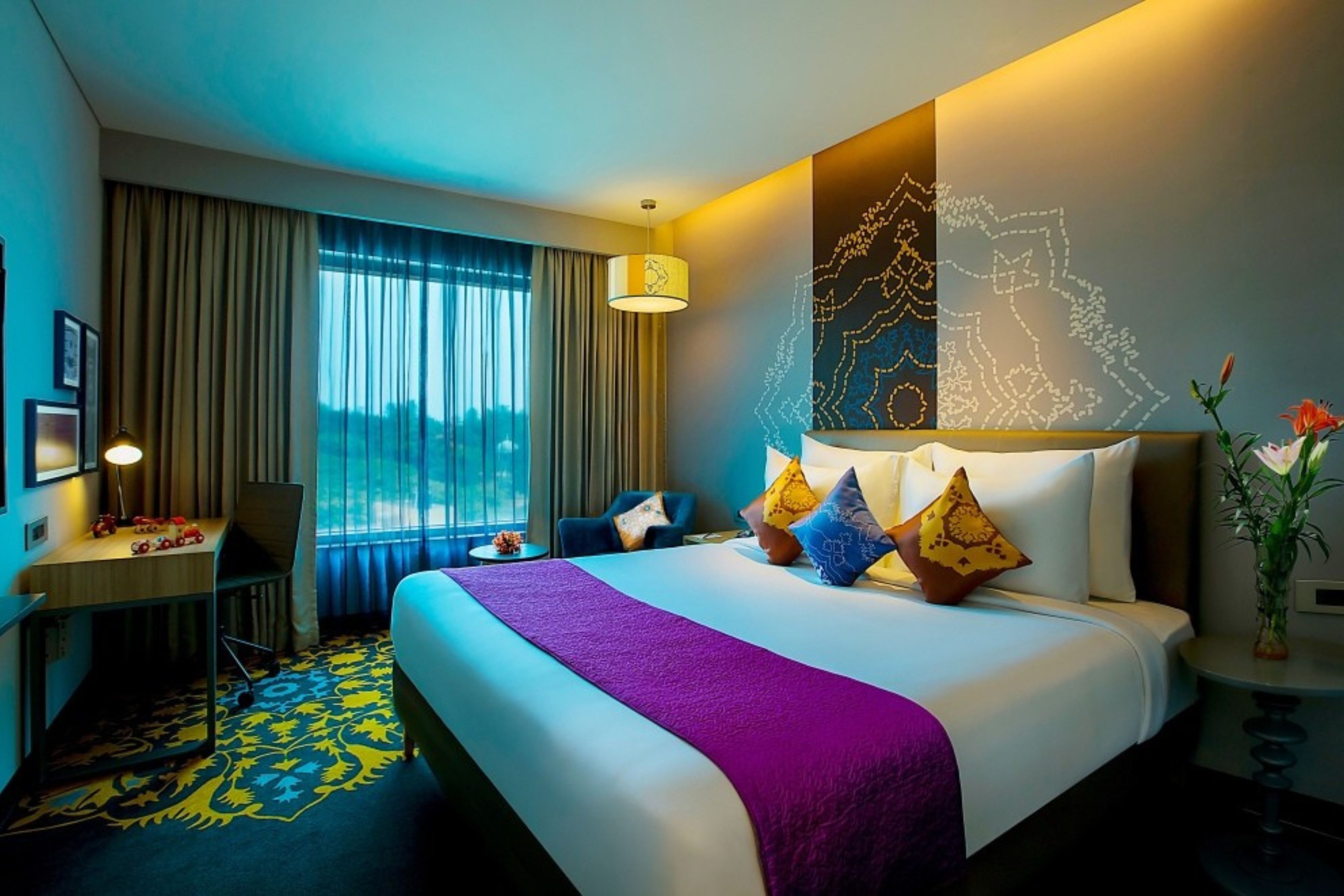 A hotel room with a bed, a desk, a chair, a lamp, and a window with curtains.