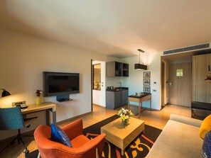 A modern living room with a flat-screen TV, a sofa, a coffee table with flowers, a desk with a lamp, and a kitchen area with cabinets and a sink.