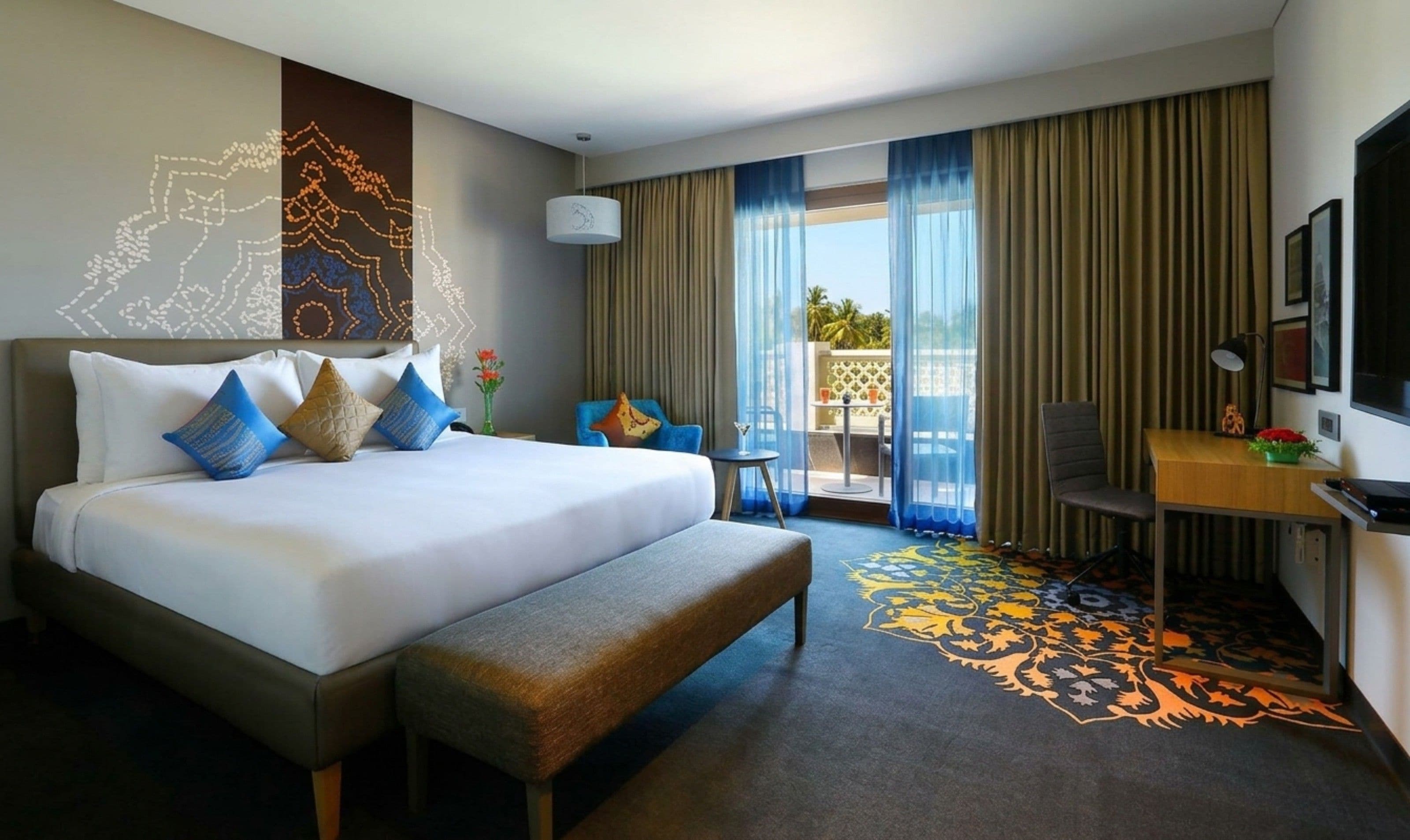 A hotel room with a large bed, a desk, a chair, a TV, and a balcony with curtains.