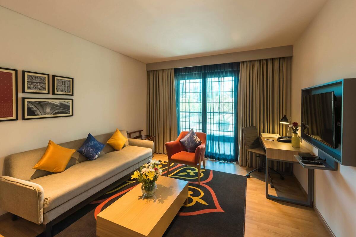 A modern hotel room with a sofa, armchair, desk, TV, and large windows.
