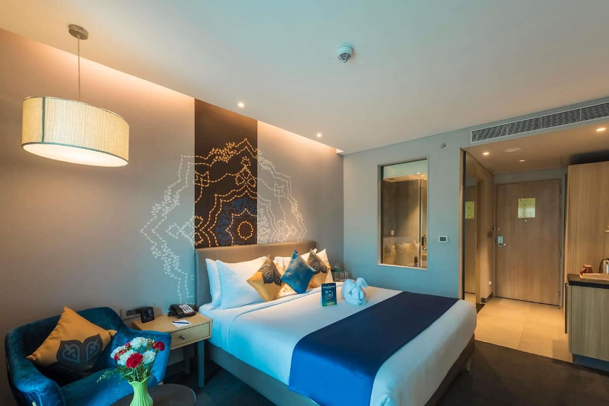 A modern hotel room with a bed, bedside table, lamp, and a seating area with a sofa and table.