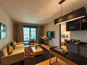 A modern hotel room with a sofa, coffee table, TV, kitchenette, and a large window with curtains.