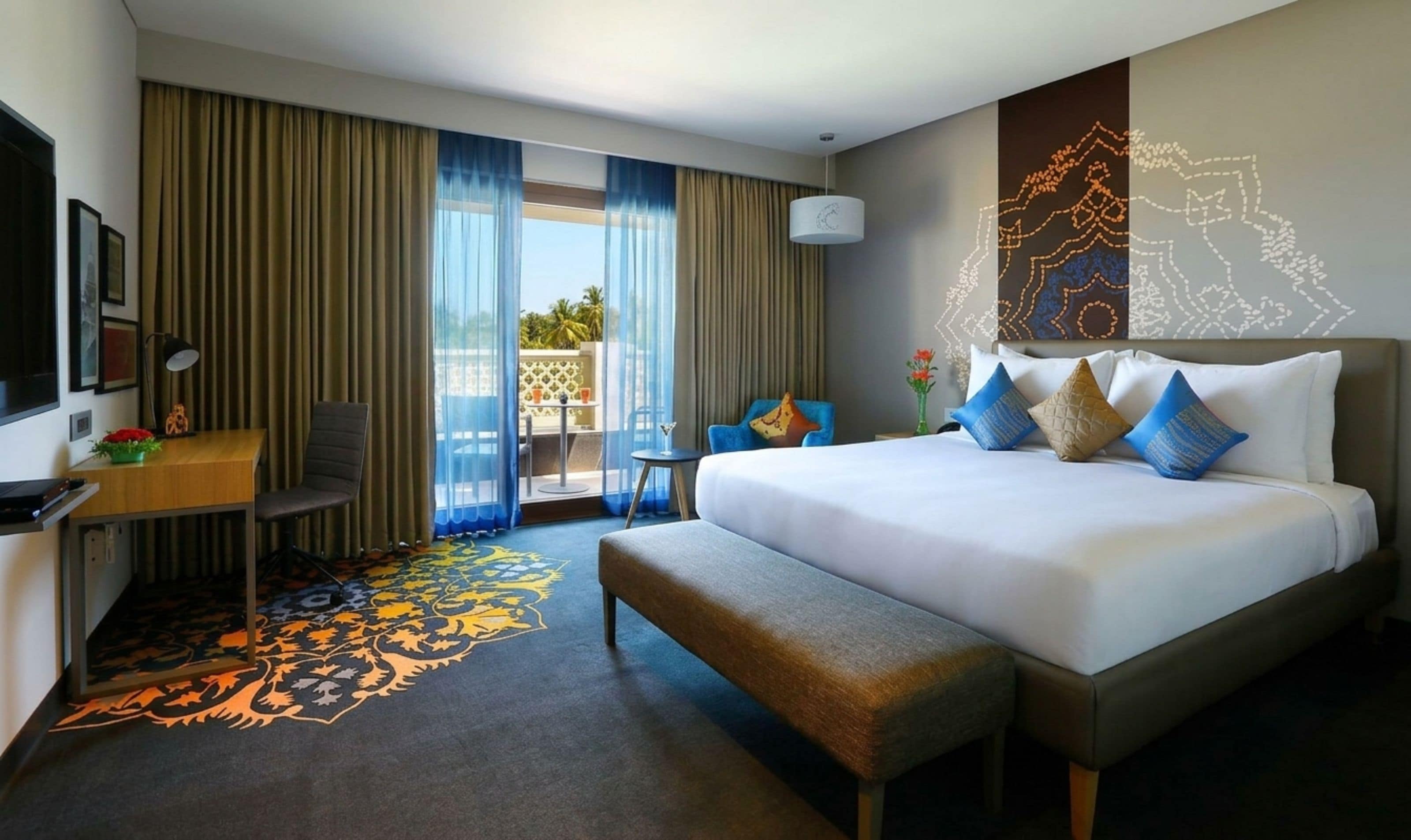 A hotel room with a bed, a desk, a chair, a bench, a TV, and a view of the outdoors.