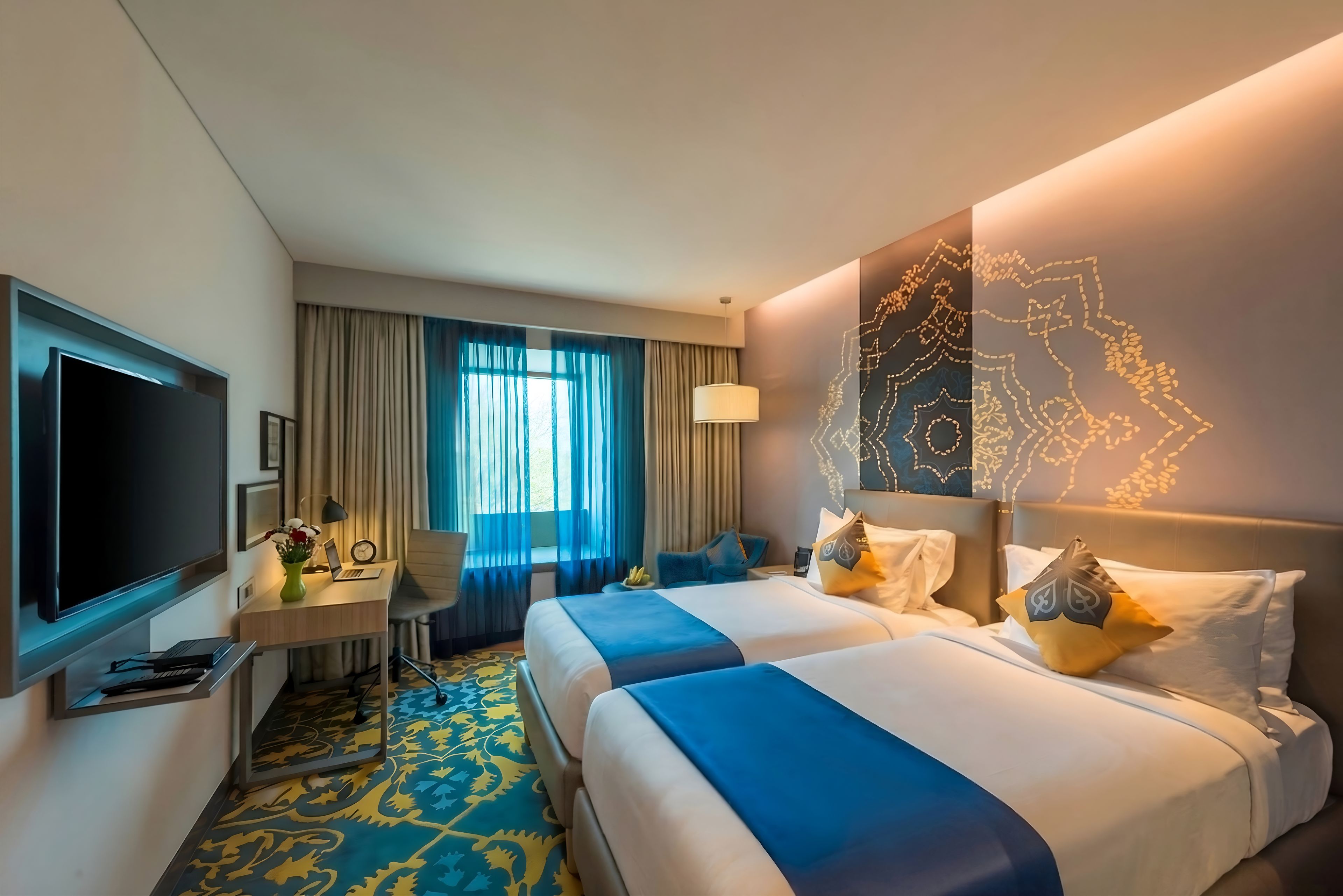 A hotel room with two beds, a desk with a laptop, a TV, and a large window with curtains.