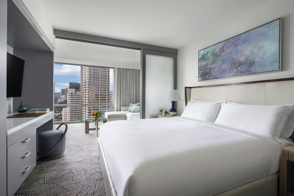 A hotel room with a large bed, a desk with a chair, a TV, and a city view through large windows.