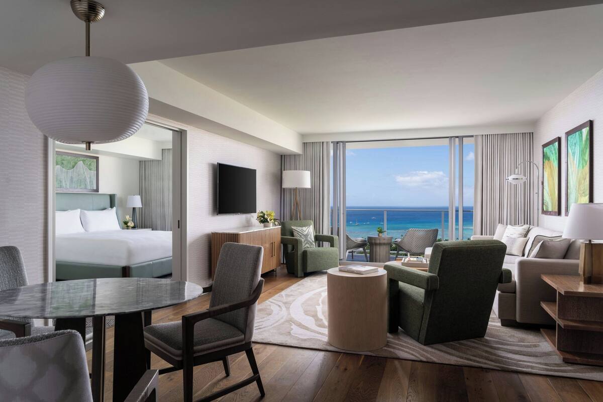 A modern hotel room with a large bed, a dining area, a seating area with a view of the ocean, and a flat-screen TV.