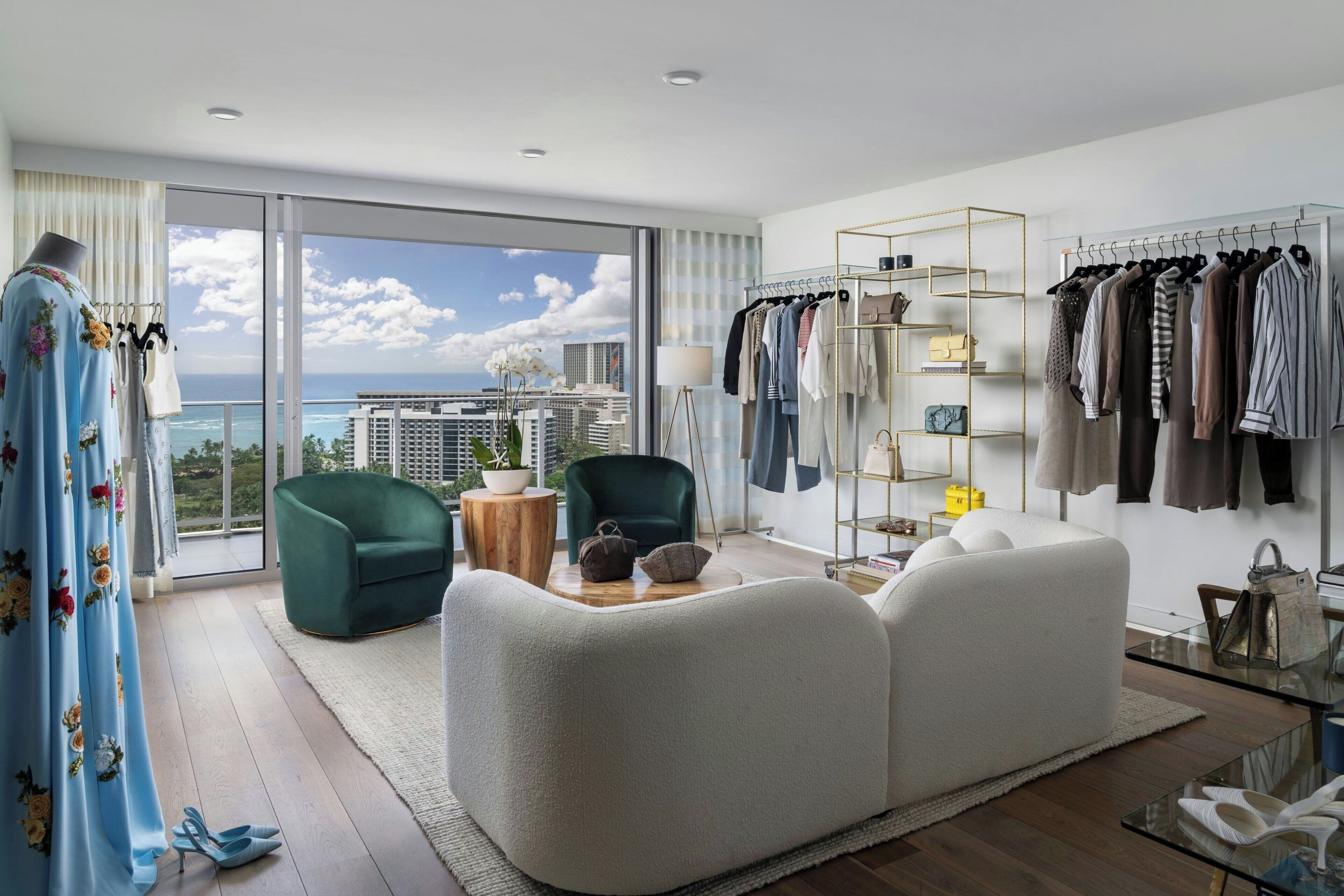 Un salon moderne doté d’une grande fenêtre, d’un fauteuil vert, d’un canapé blanc et d’un portant à vêtements sur lequel sont disposés différents articles.