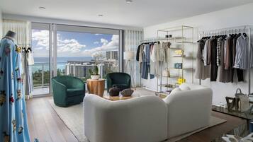 A modern living room with a large window, a green armchair, a white sofa, and a clothing rack with various garments.