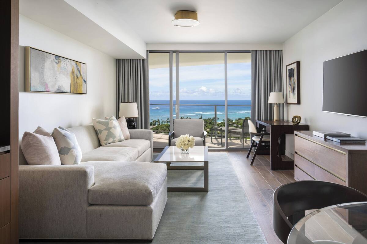 A modern hotel room with a large balcony, a sofa, a coffee table, and a TV.
