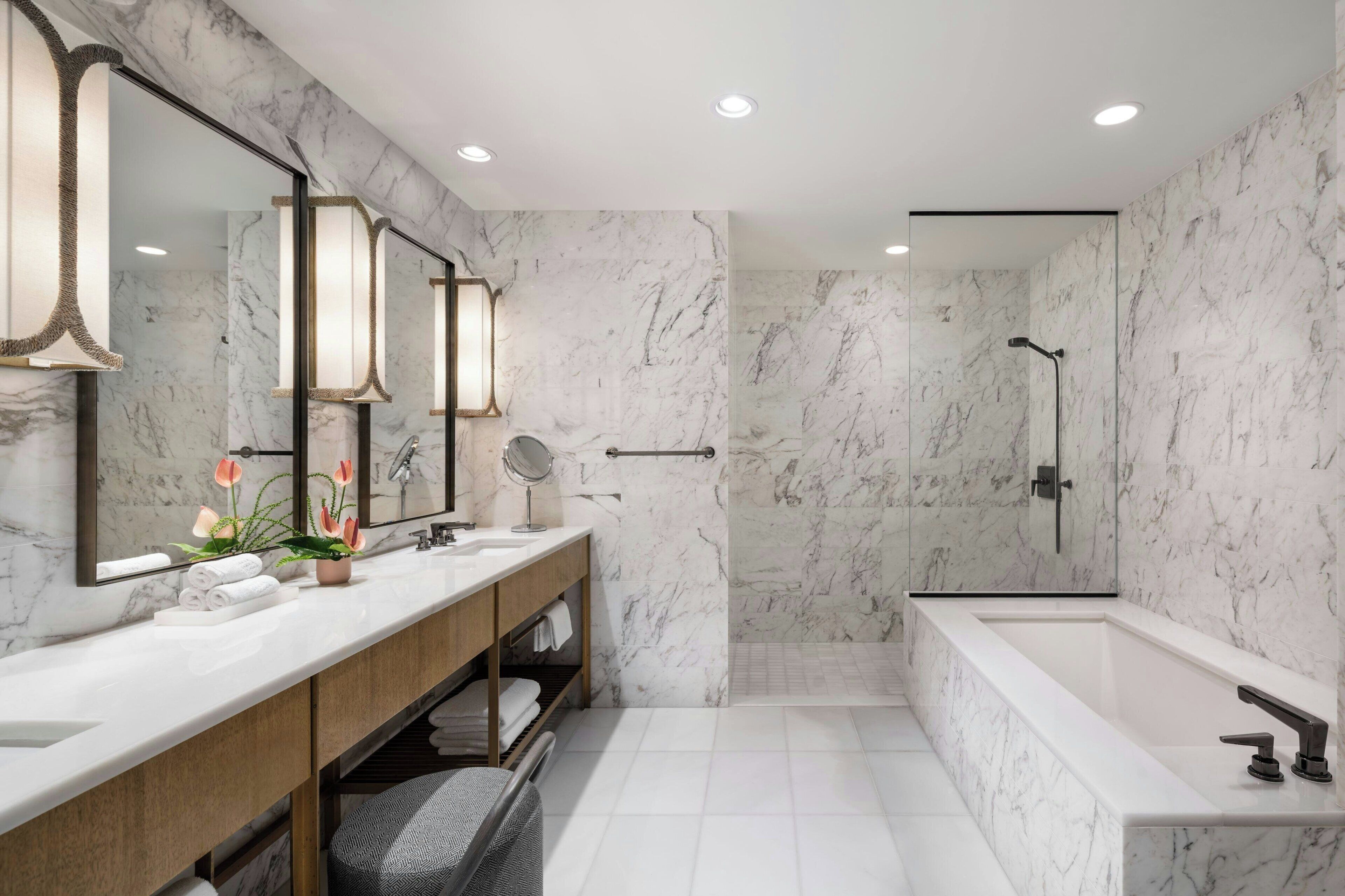 A modern bathroom with marble walls, a large bathtub, a shower, and a vanity with a mirror and towel rack.