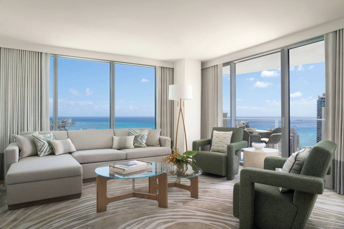 A modern living room with a large sofa, armchairs, a glass coffee table, and a view of the ocean.
