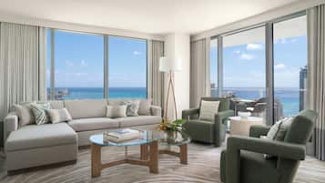A modern living room with a large sofa, armchairs, a glass coffee table, and a view of the ocean.