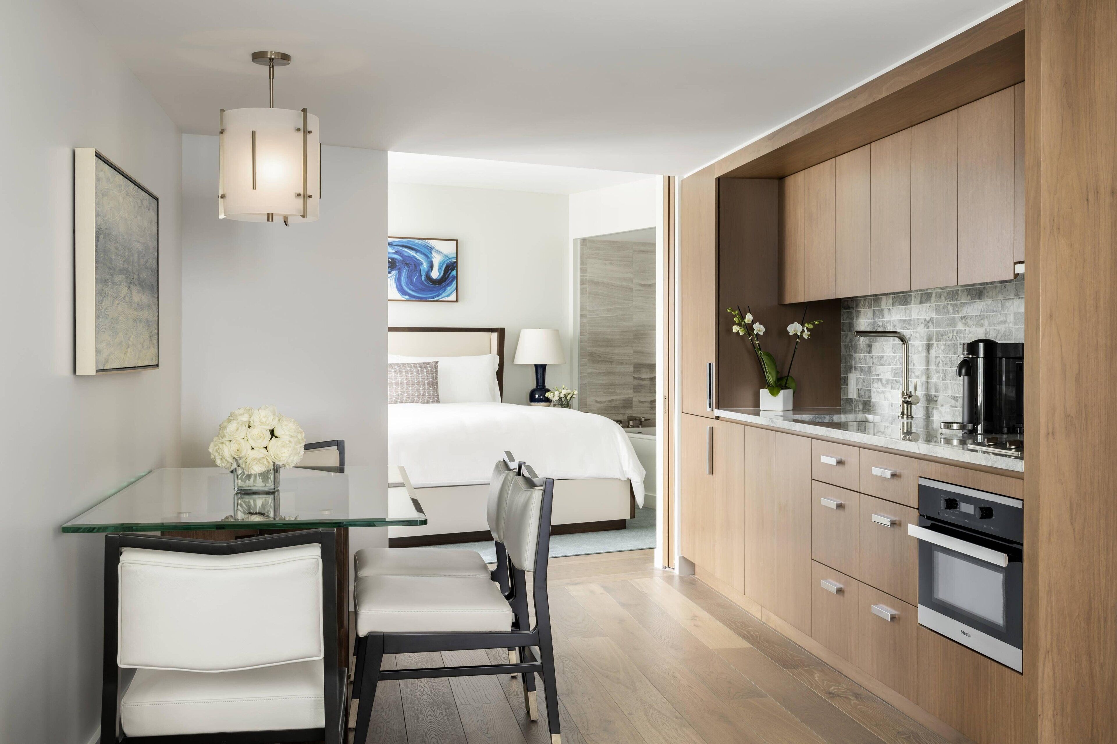 A modern hotel room with a glass dining table, a bed with white linens, a kitchenette with wooden cabinets, and a large window.