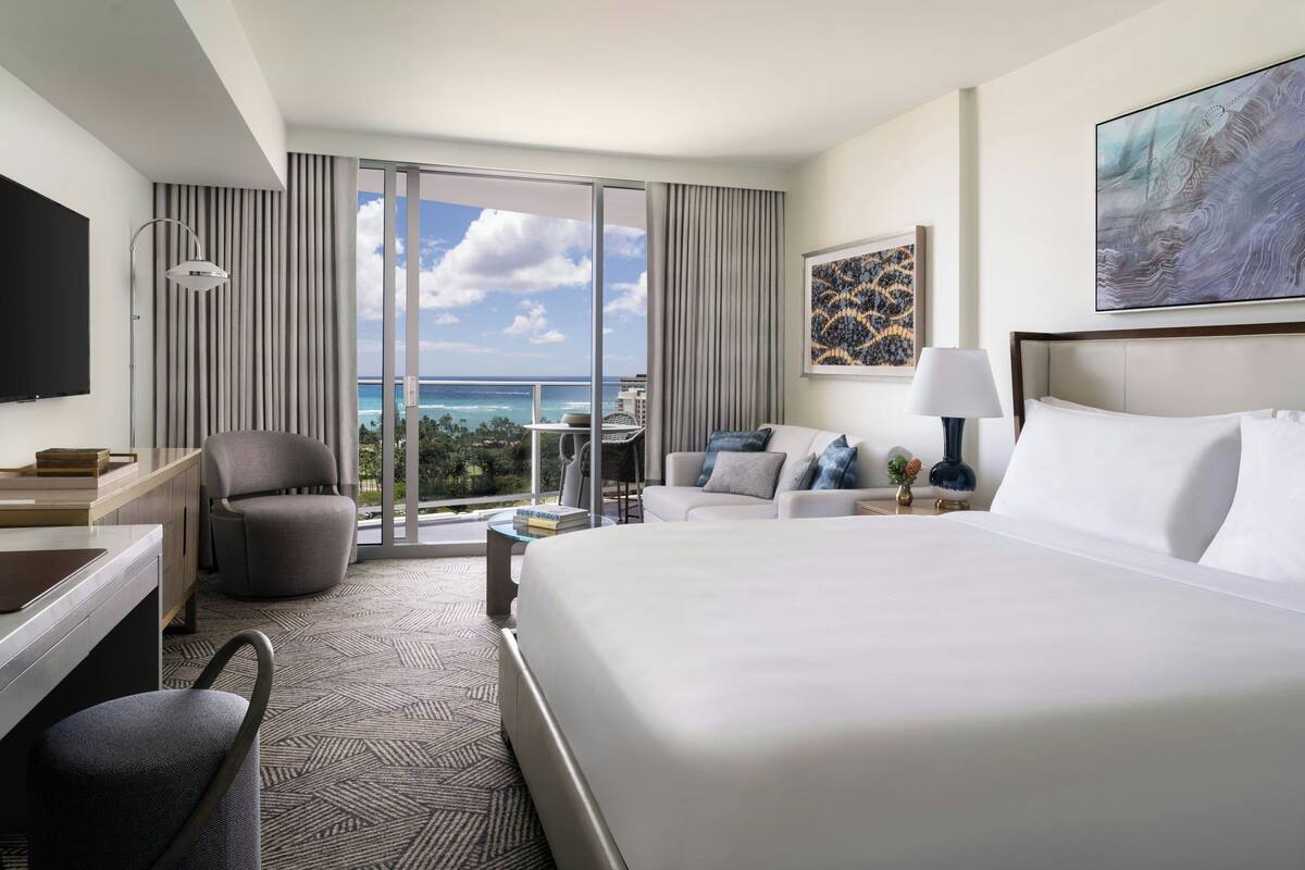 A hotel room with a large bed, a desk, a chair, a sofa, and a TV.