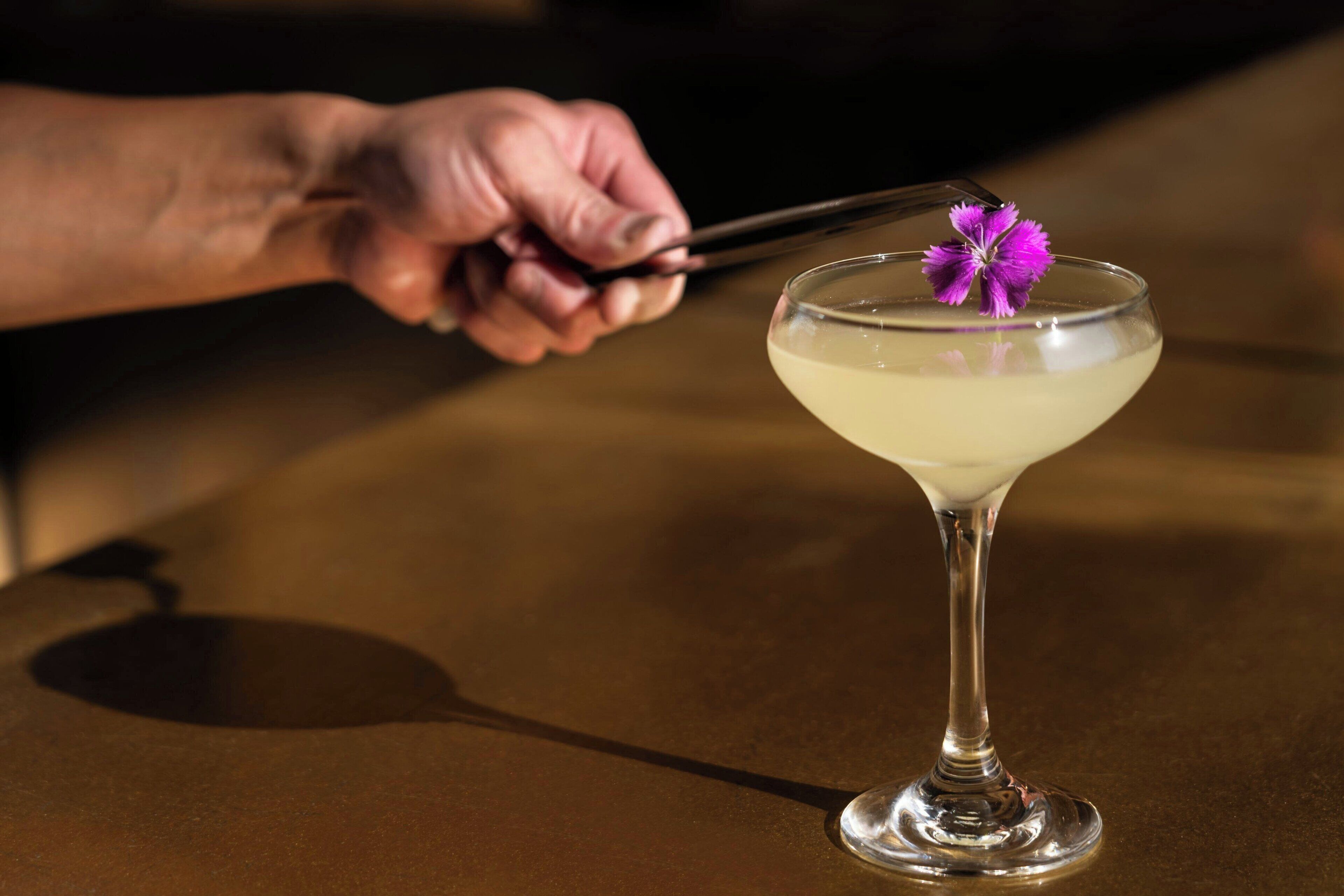 A cocktail in a martini glass with a purple flower garnish, on a table.