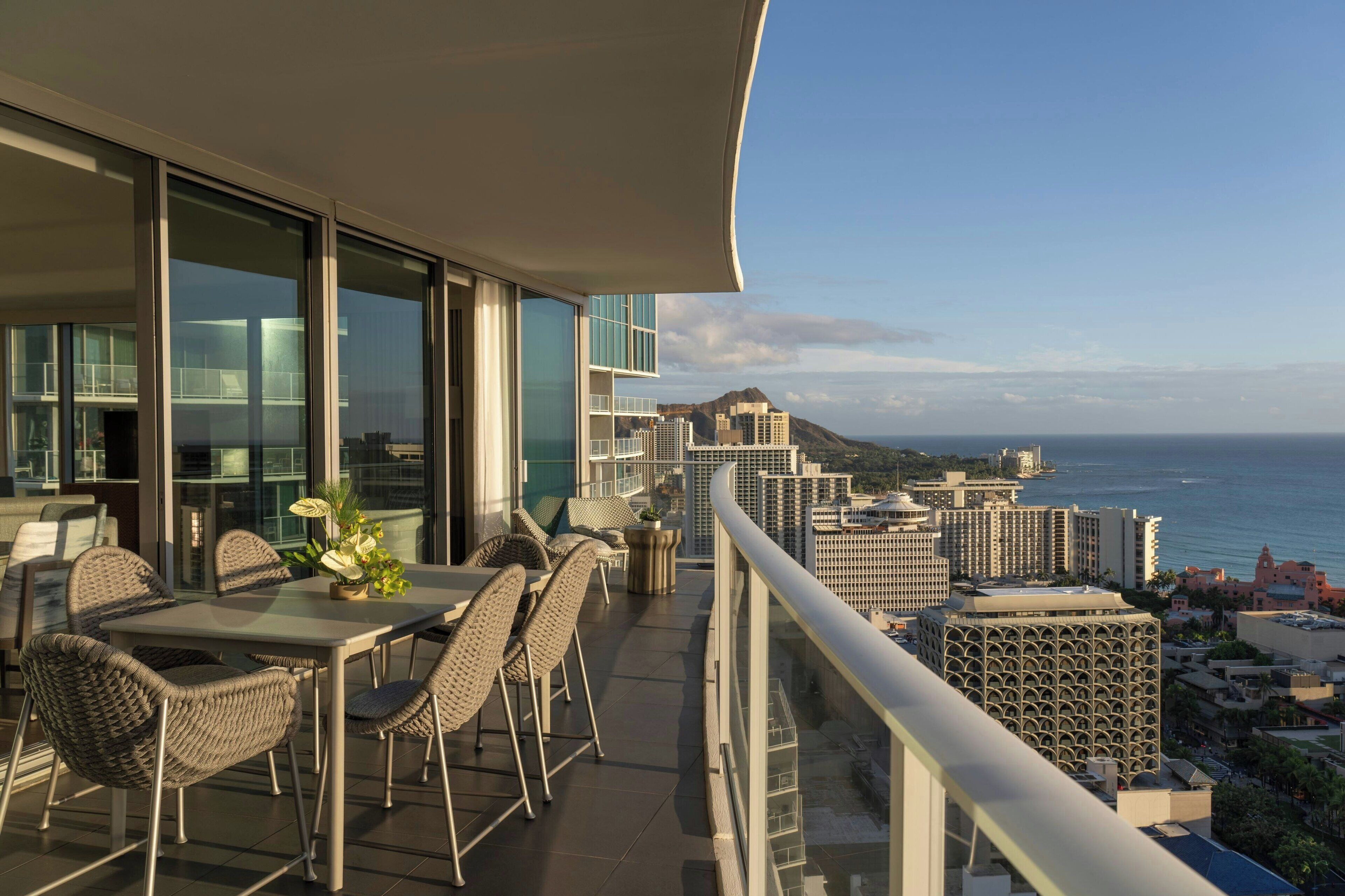 A balcony with a dining area, large windows, and a view of the ocean and cityscape.