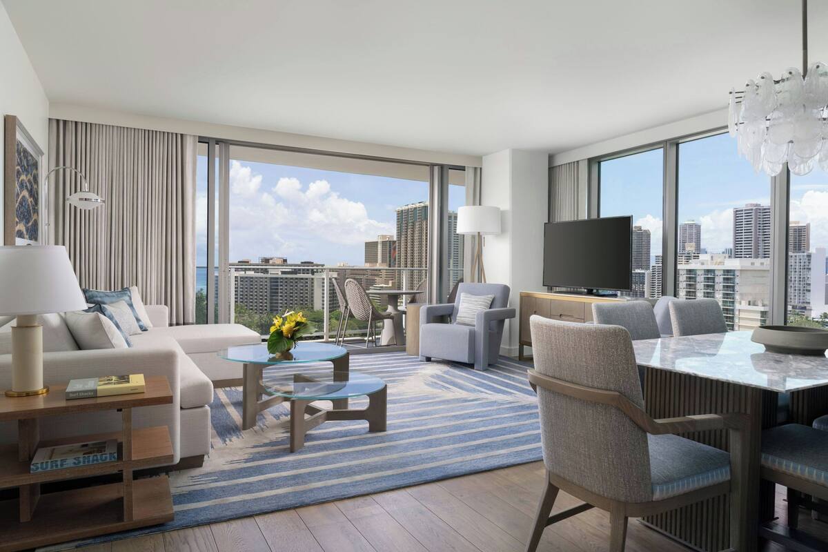 A modern hotel room with a large balcony, a sofa, a dining table, and a TV.