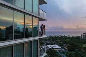 A high-rise building with large windows and a balcony overlooking a cityscape and ocean.