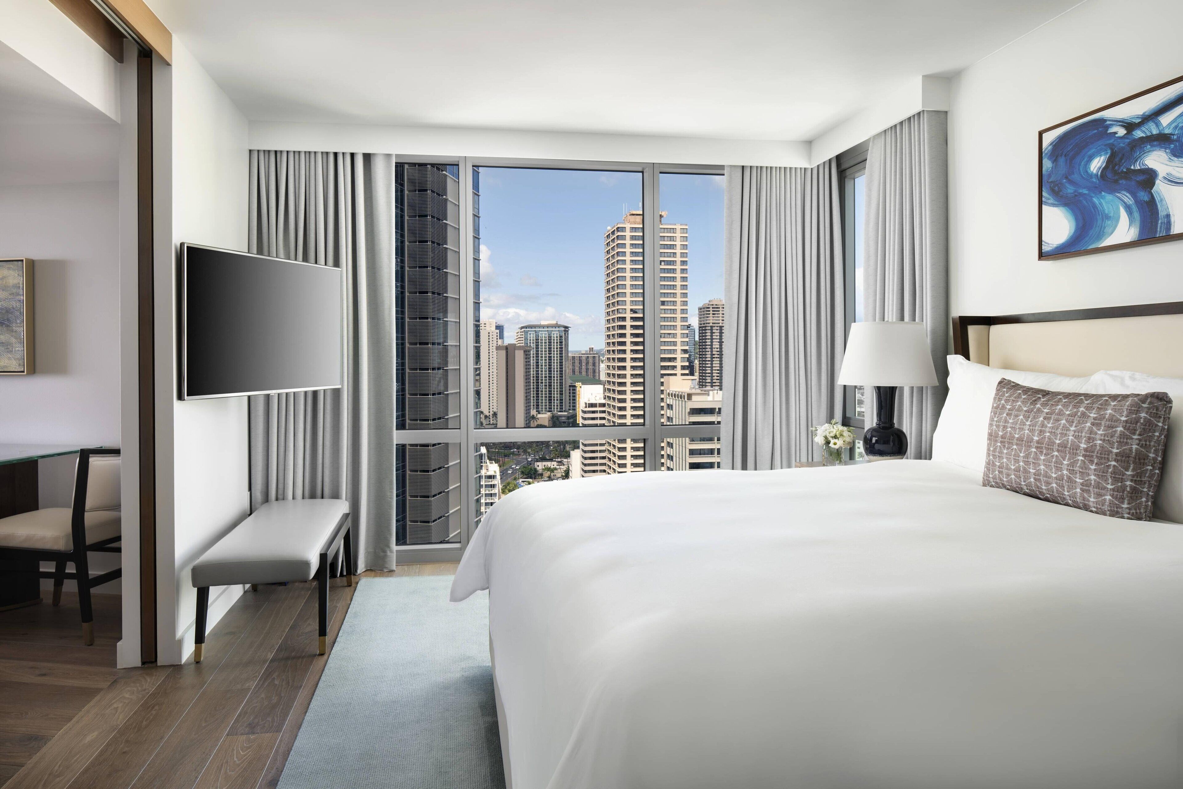 A hotel room with a large bed, a flat-screen TV, a bench, and a city view through floor-to-ceiling windows.