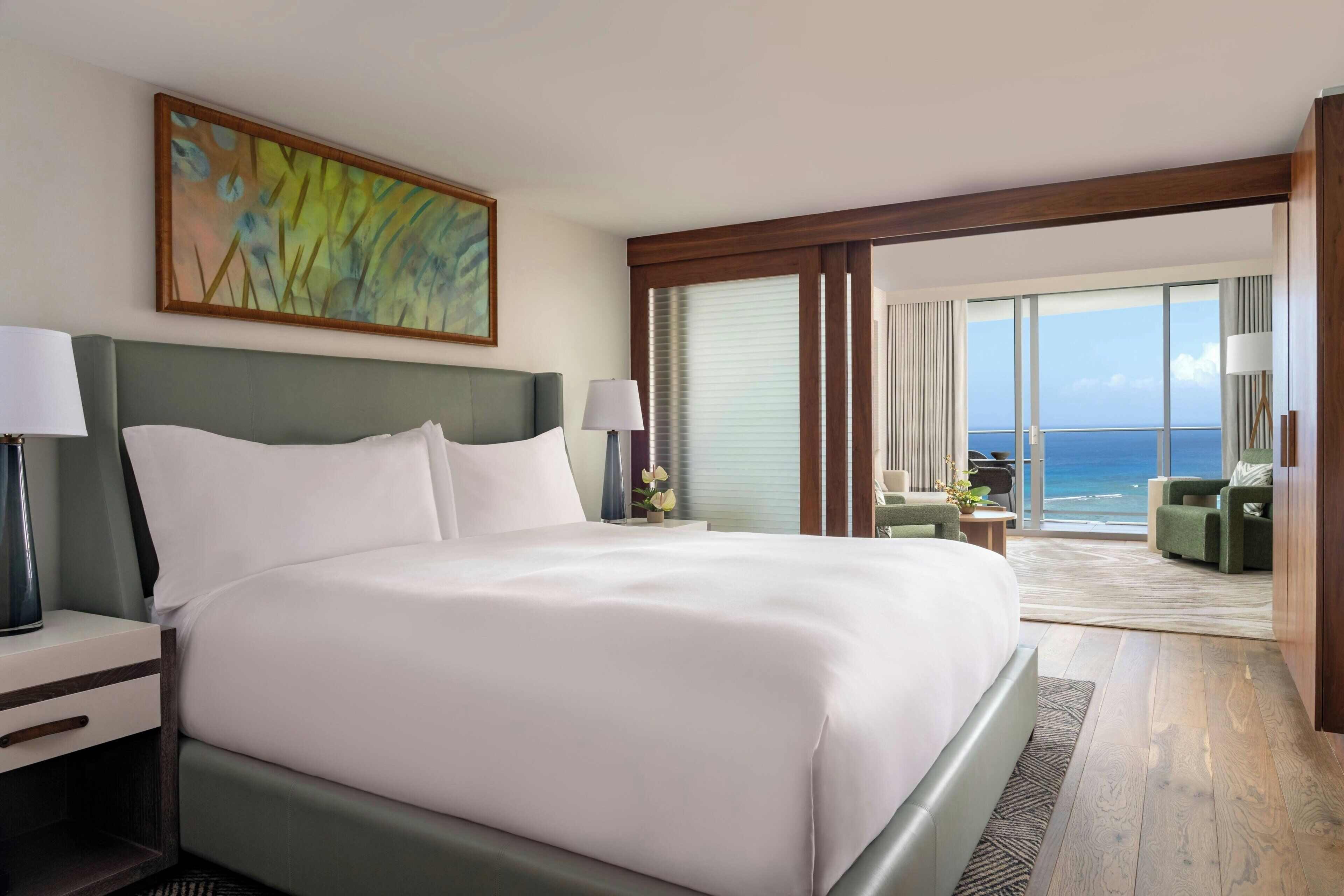 A large bed with a headboard, two bedside tables with lamps, a painting above the bed, and a view of the ocean through large windows.
