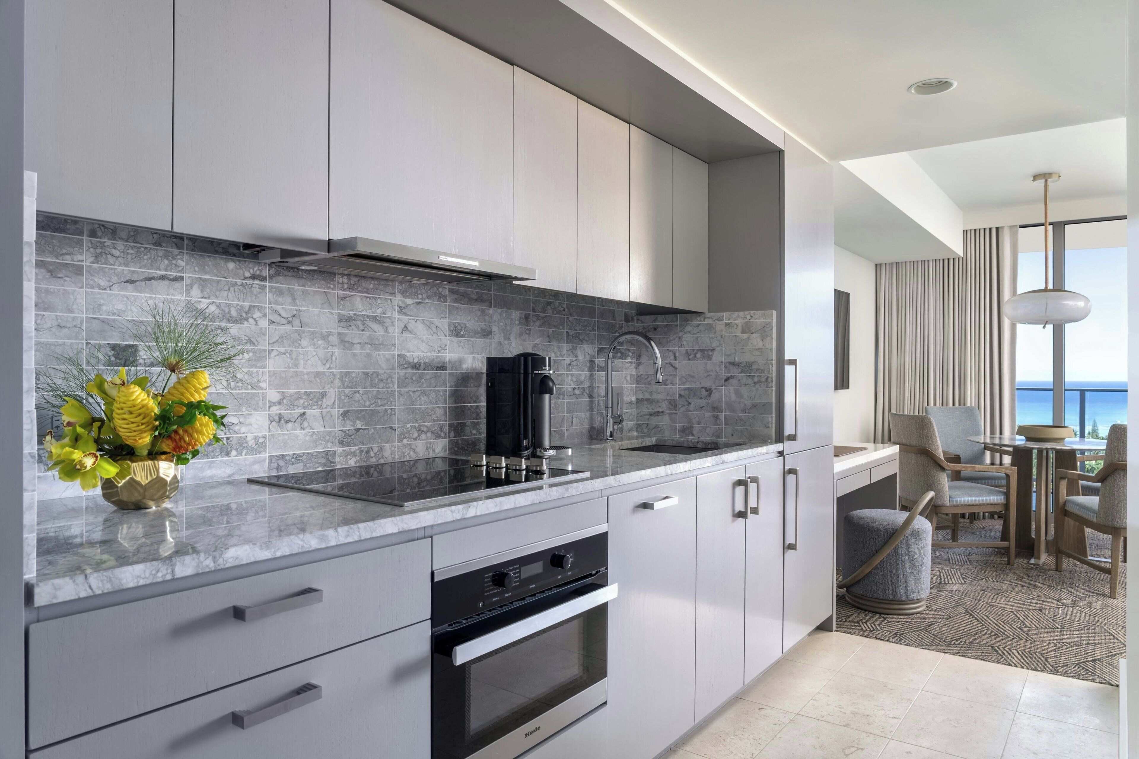 A modern kitchen with grey cabinets, a marble backsplash, and a coffee machine. There is a dining area with a table and chairs, and a view of the ocean through large windows.