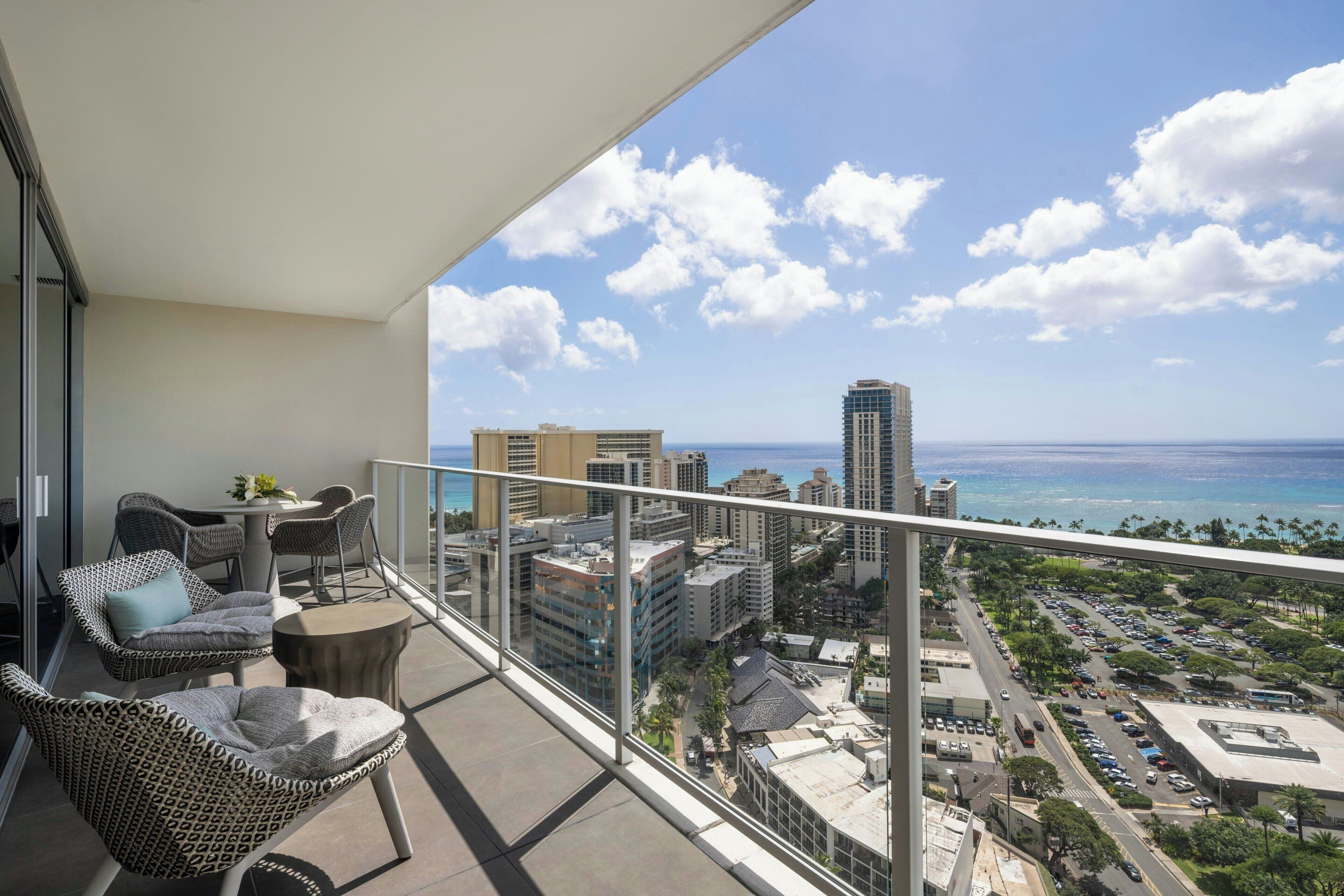 A balcony with wicker chairs and a table, overlooking a cityscape with buildings and a view of the ocean.
