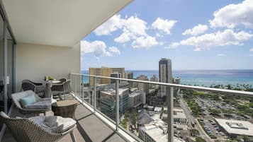 A balcony with wicker chairs and a table, overlooking a cityscape with buildings and a view of the ocean.