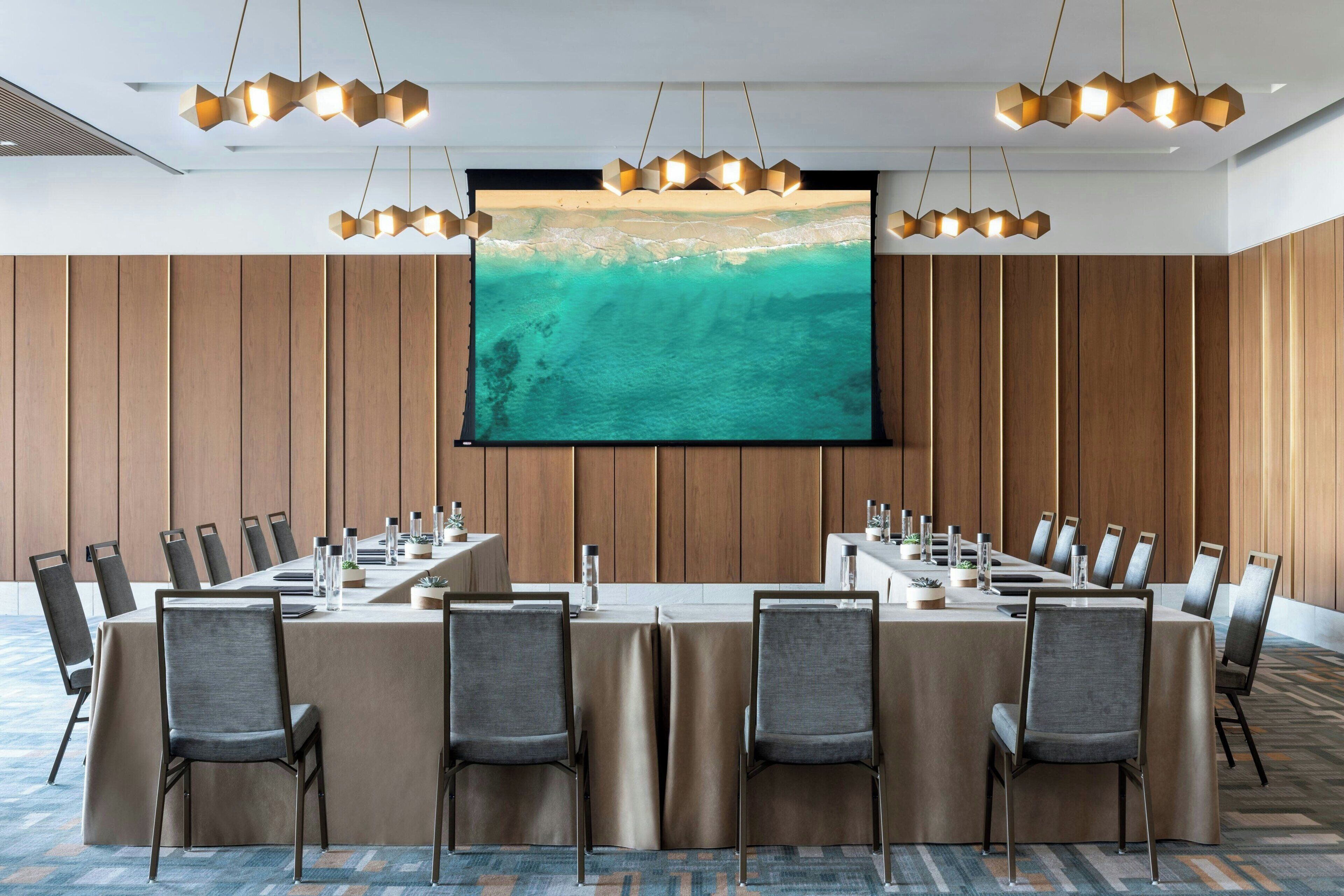 Un salón de conferencias con una larga mesa, varias sillas y una pantalla grande que muestra una escena de playa.