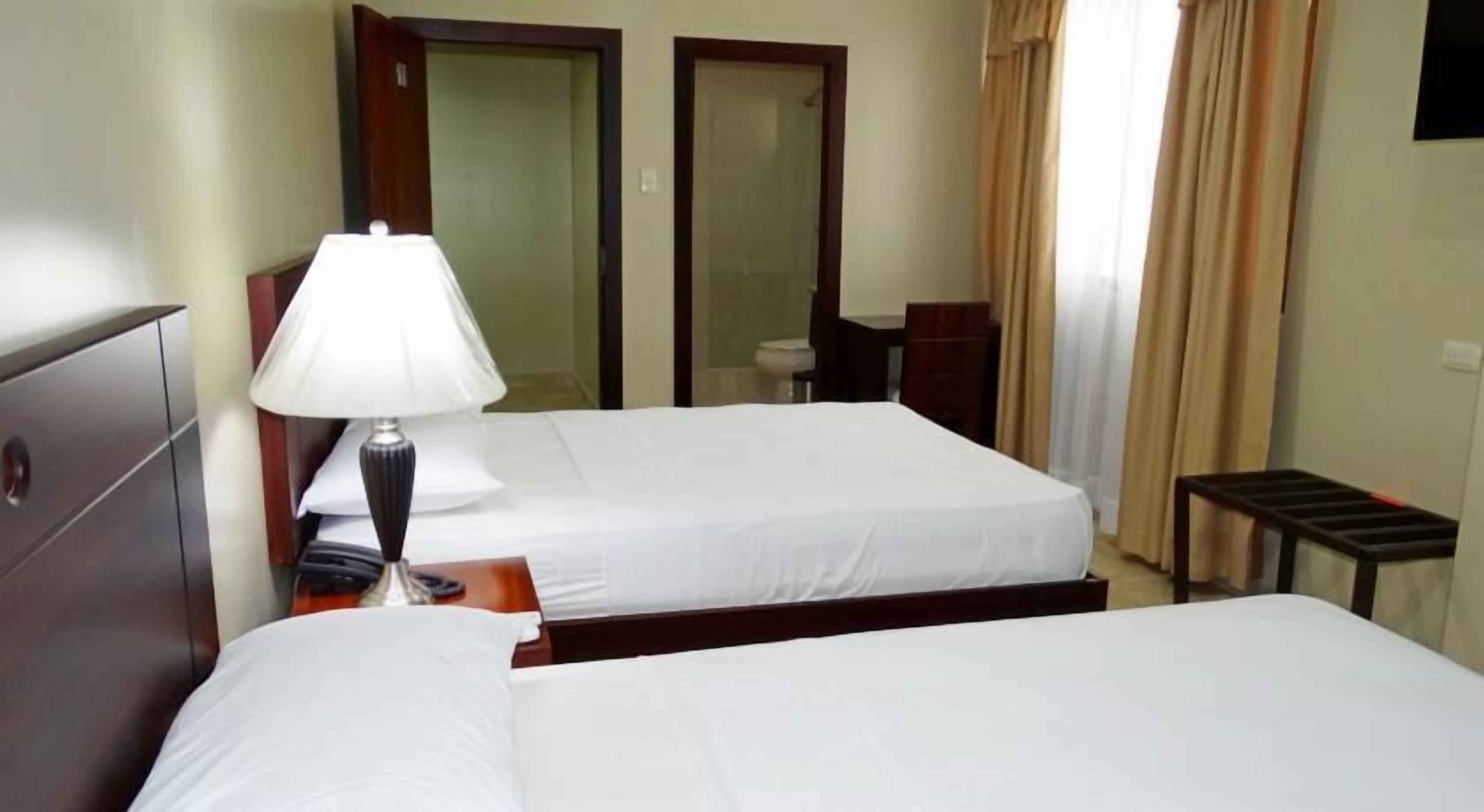 A hotel room with two single beds, a bedside table with a lamp, a wooden headboard, a mirror, a small desk, and a window with curtains.