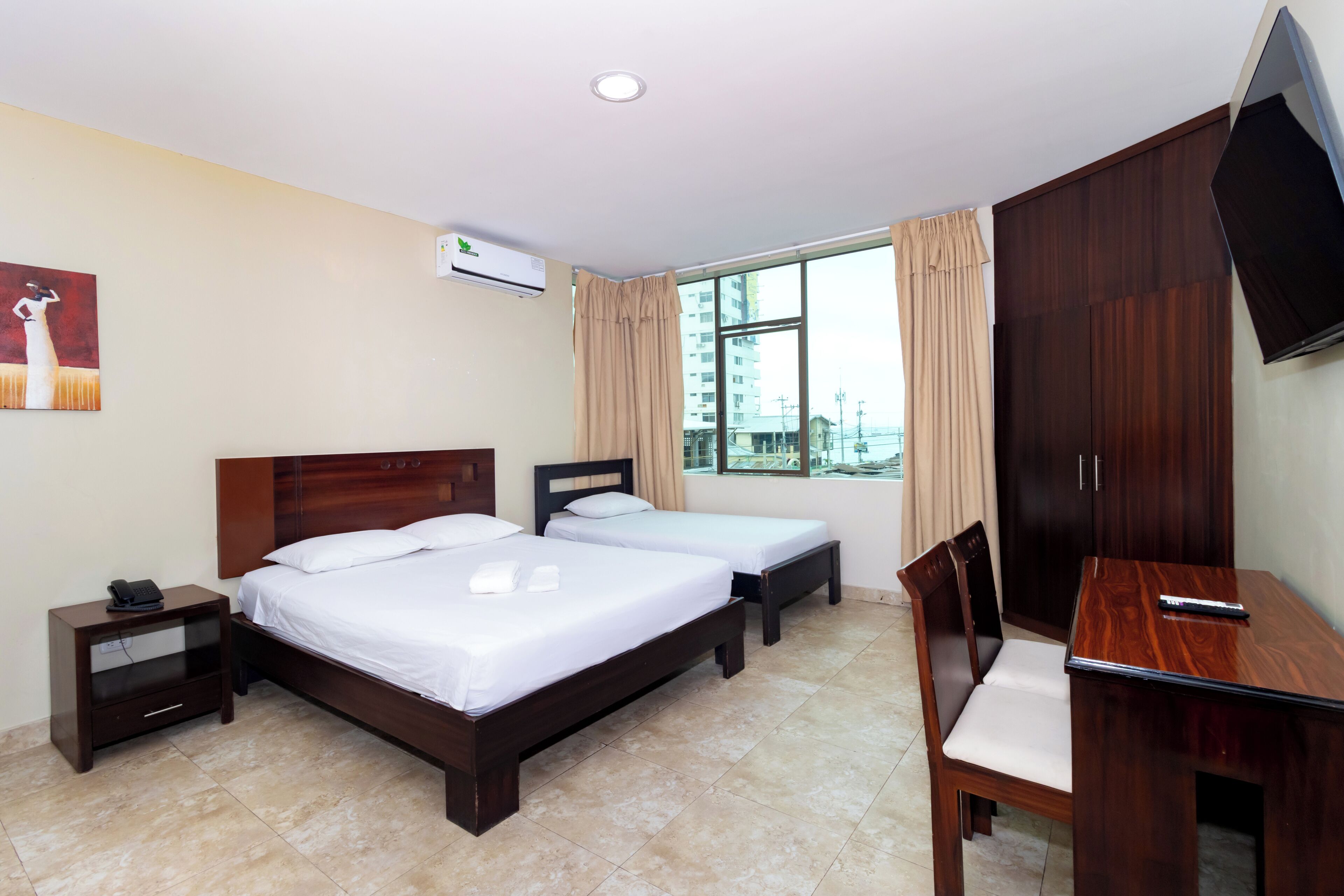 A hotel room with two beds, a desk, a chair, a TV, an air conditioner, and a window with curtains.
