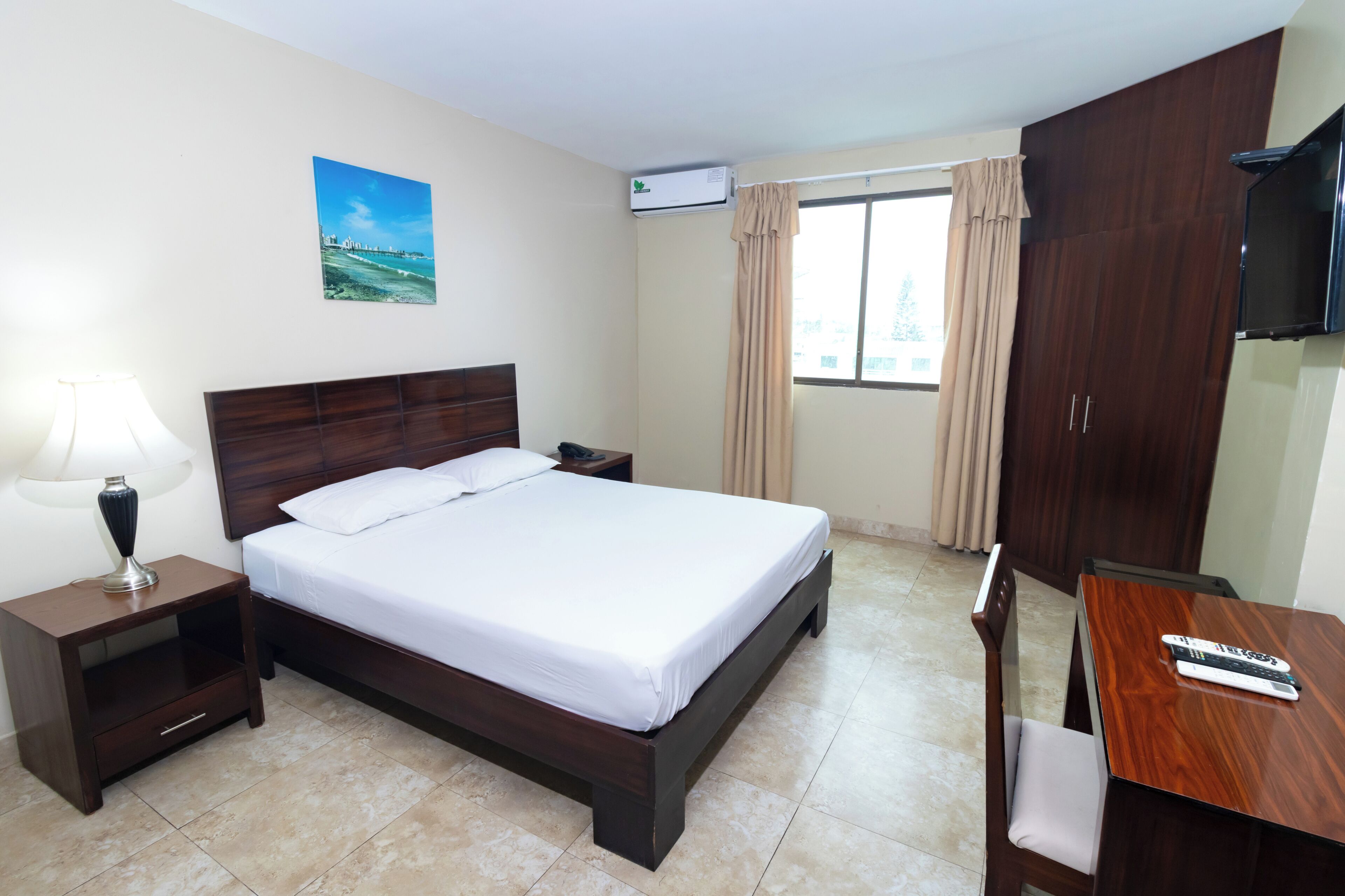 A hotel room with a bed, bedside table, lamp, TV, and a view of the outdoors.