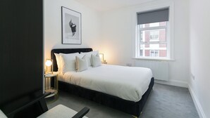 Un dormitorio con cama, lámparas de noche, un cuadro en la pared y una ventana con vistas a edificios.