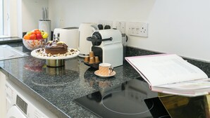 Una encimera de cocina con cafetera, un pastel, un frutero y un libro.
