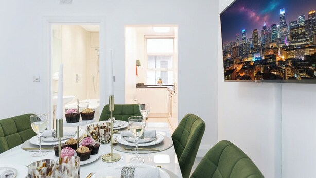 Un área de comedor con vista a la ciudad, sillas tapizadas de verde y una mesa blanca con platos, vasos y cupcakes.