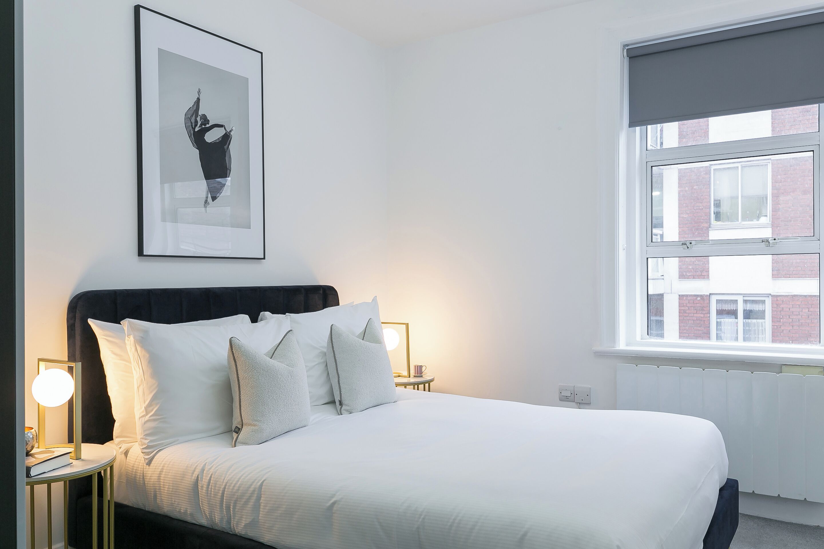Een net opgemaakt bed met witte lakens, een lijst met een afbeelding erboven en nachtlampjes op een nachtkastje.