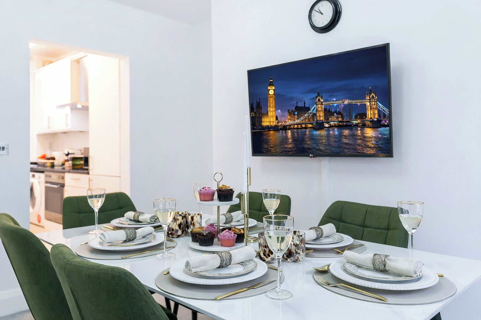 A dining area with a table set for four, featuring white plates, gold cutlery, and green chairs. A painting of a cityscape with a bridge and tower is on the wall.