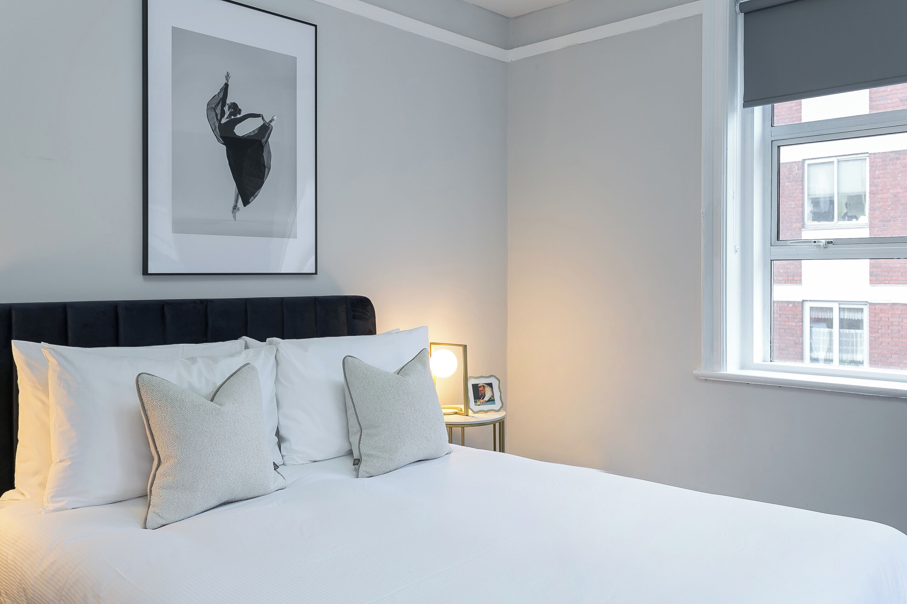 A neatly made bed with white linens and two pillows, a framed picture on a bedside table, and a window with a view of a brick building.