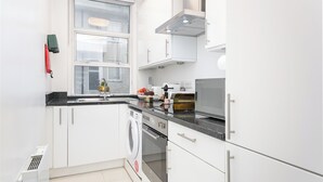 Una cocina moderna con armarios blancos, horno empotrado, lavadora y ventana con persianas.