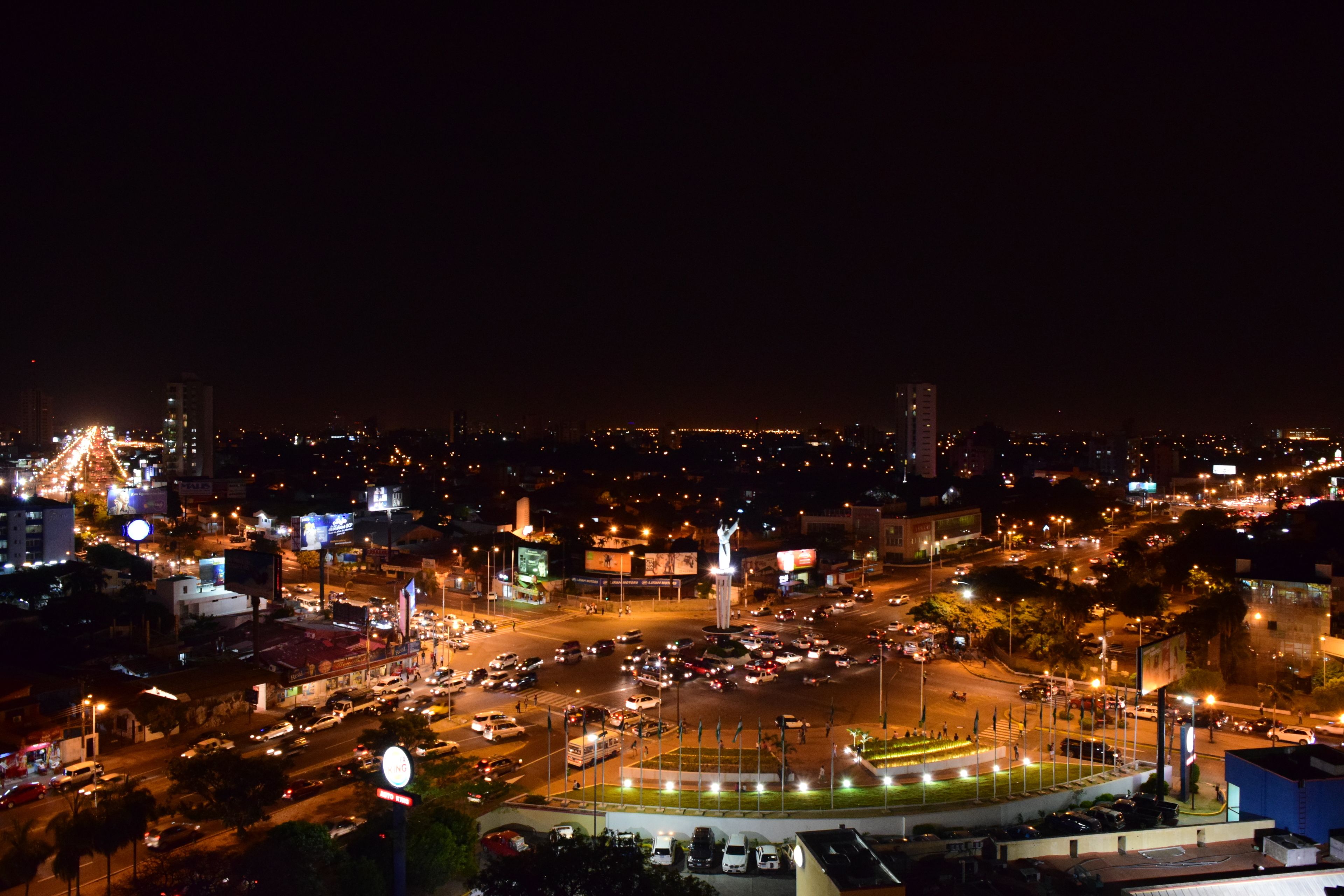A nighttime cityscape with a central roundabout, illuminated buildings, and a statue.
