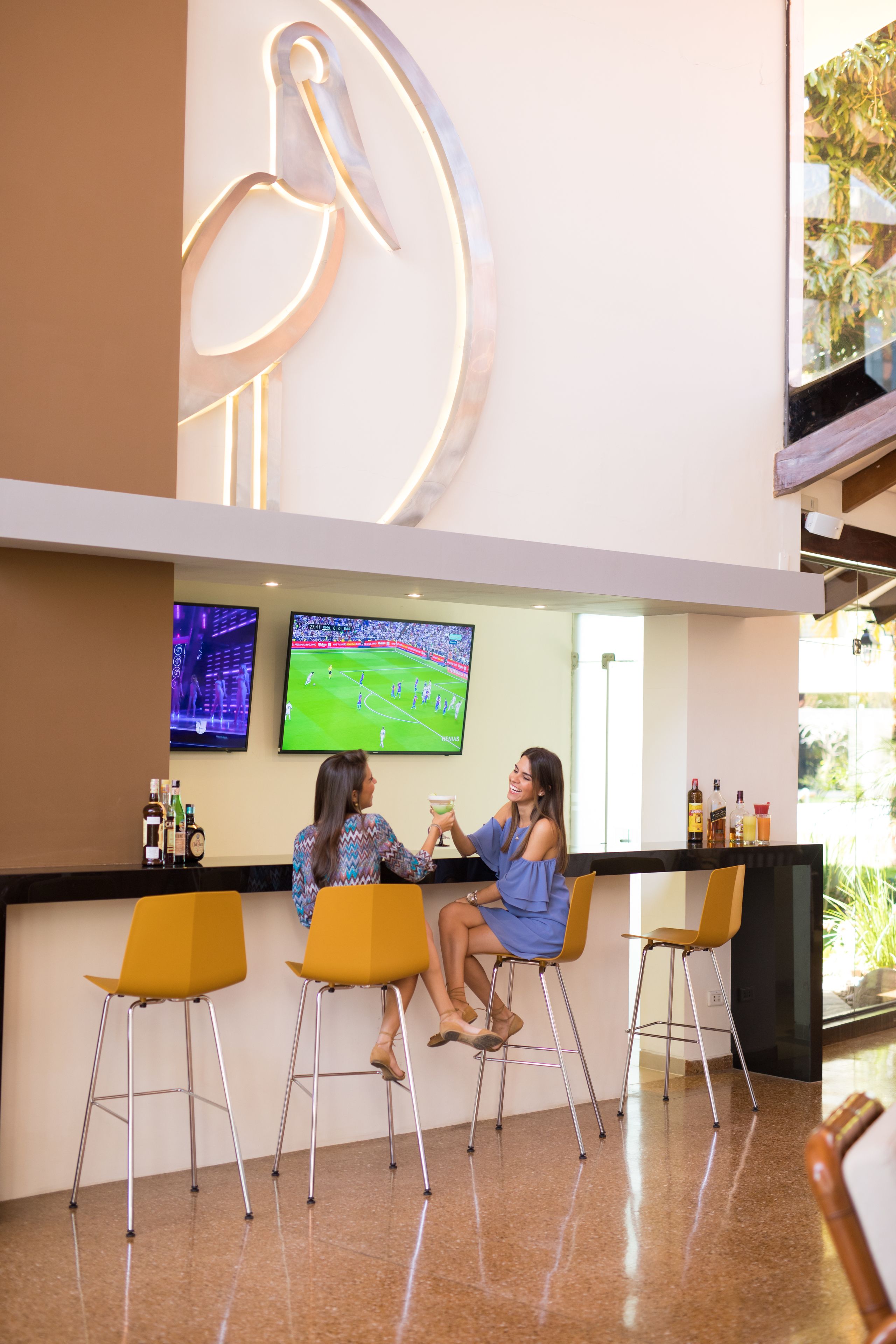 Una zona de bar con tres sillas amarillas, una pantalla grande montada en la pared mostrando un evento deportivo y una pieza decorativa en la pared.