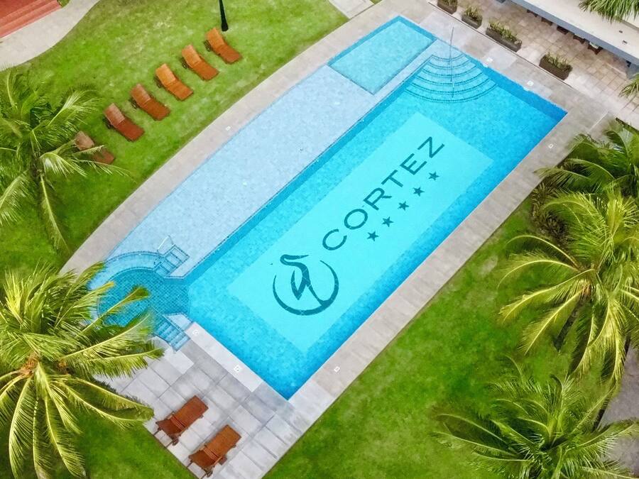 An aerial view of a swimming pool with the name 'CORTENZ' and a logo, surrounded by palm trees and a building in the background.