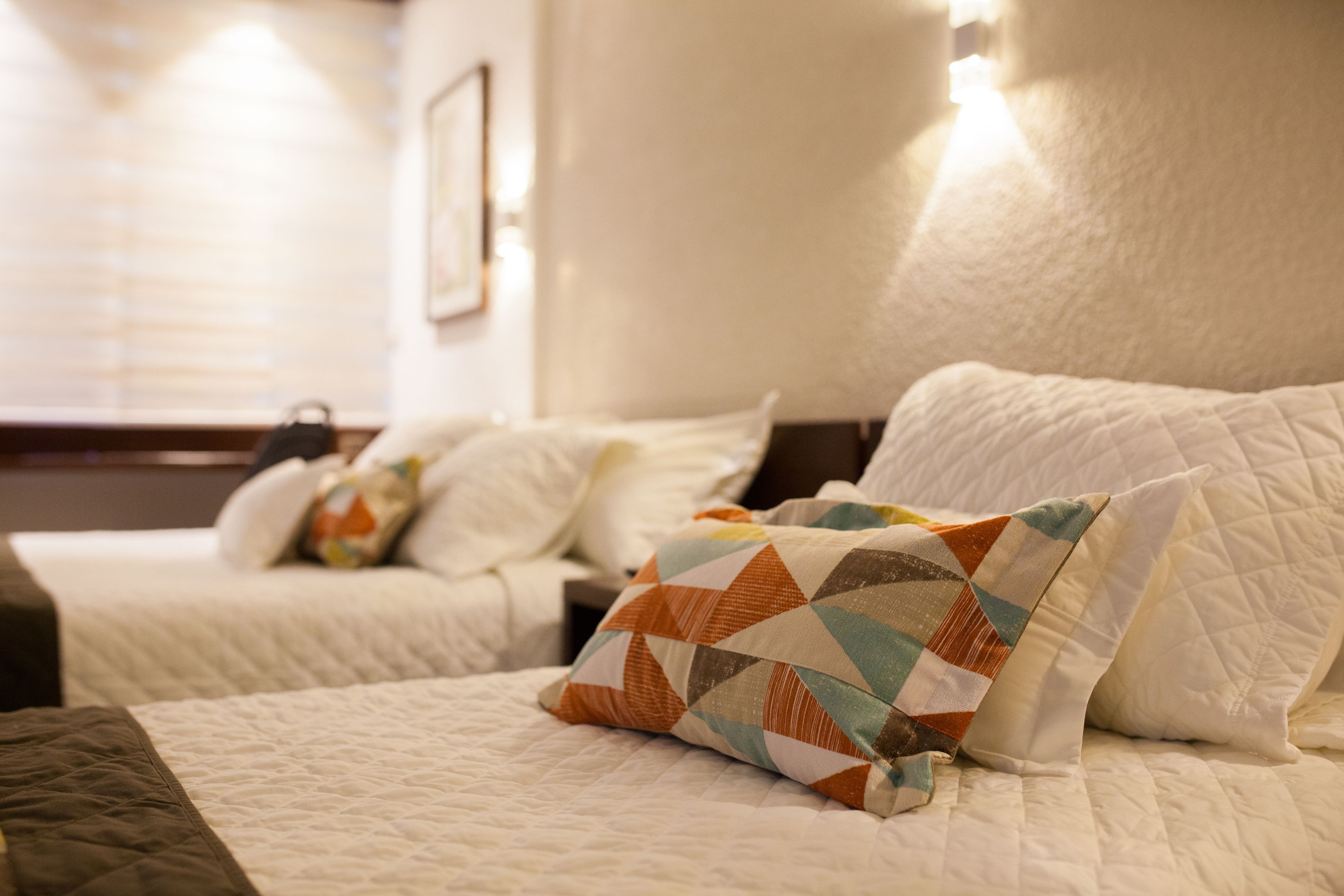 Una habitación de hotel con dos camas, cada una con ropa de cama blanca y un patrón acolchado. Una cama tiene una almohada estampada y la otra, una almohada lisa. La habitación tiene una decoración en tonos neutros, con un cabecero y una pared de color claro.