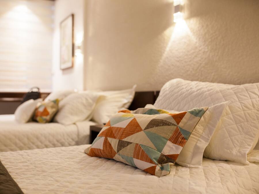 Una habitación de hotel con dos camas, cada una con ropa de cama blanca y un patrón acolchado. Una cama tiene una almohada estampada y la otra, una almohada lisa. La habitación tiene una decoración en tonos neutros, con un cabecero y una pared de color claro.