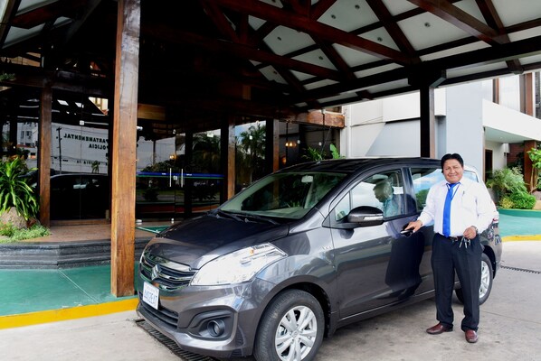 A man standing next to a car in front of a building with a covered entrance and a large billboard.