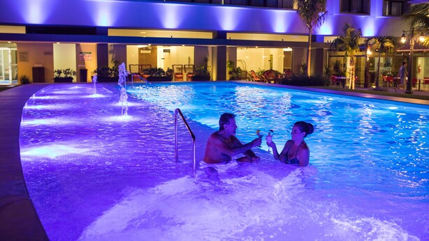 Une piscine avec un toboggan, entourée d’un bâtiment d’hôtel à plusieurs étages et dotée d’espaces de détente en plein air.