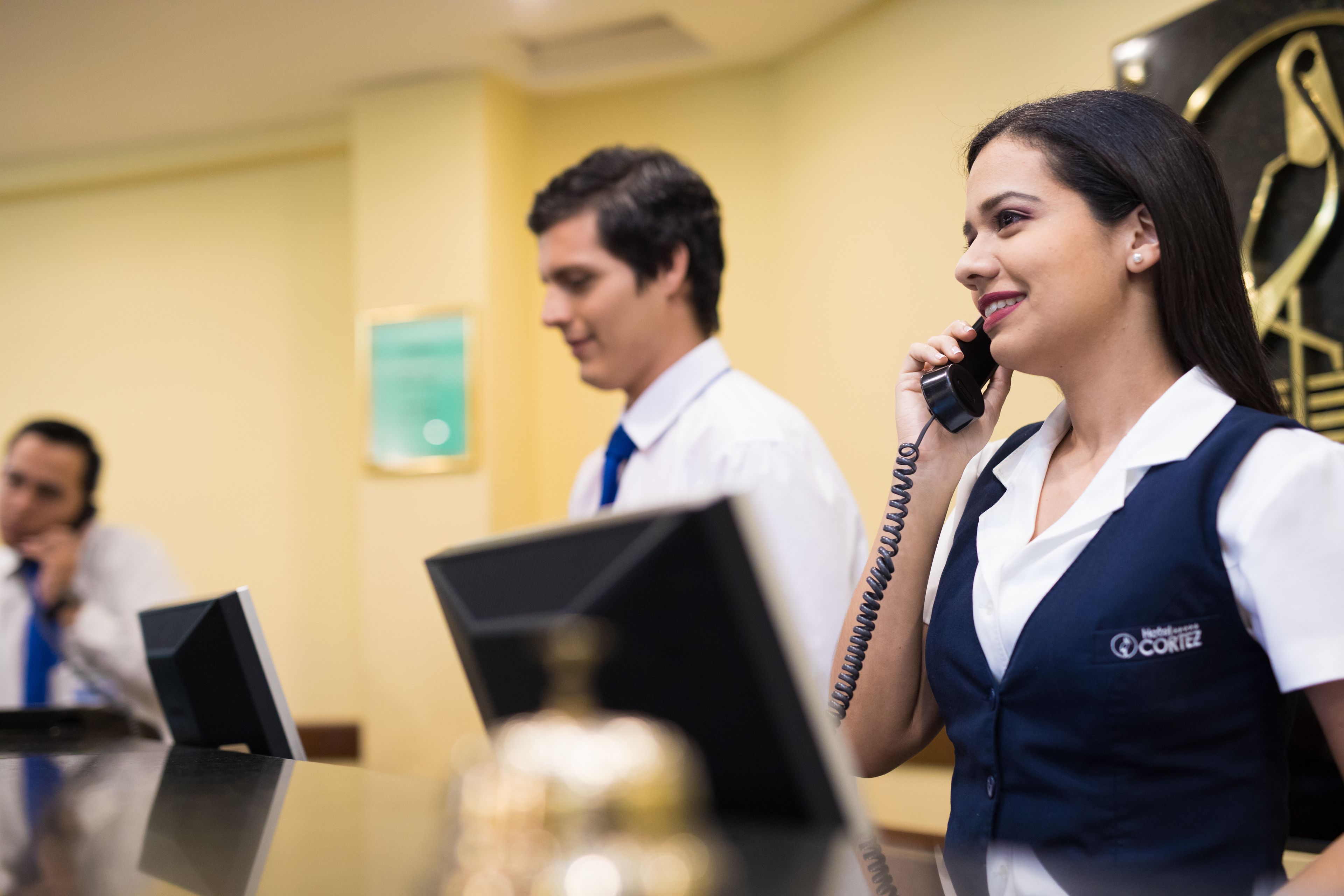 A hotel reception area with a receptionist and a guest. There are desks with computers and a clock on the wall.